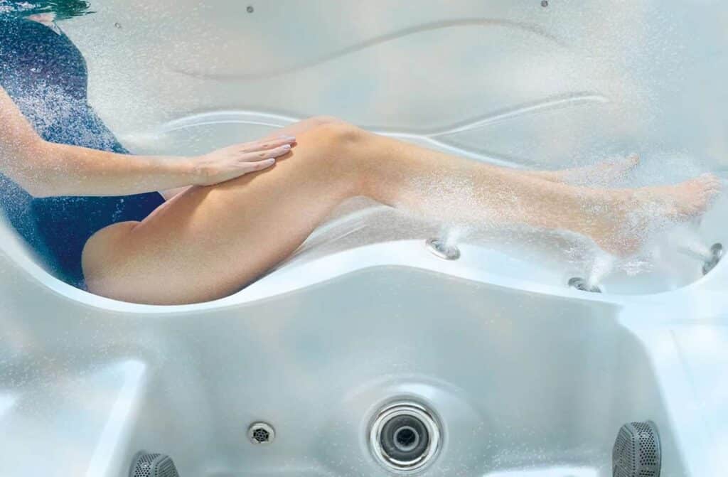 Une personne en maillot de bain bleu se détend sur un siège de salon blanc sous l'eau, avec des bulles s'élevant des jets autour de ses jambes à l'intérieur d'un spa ou d'une baignoire d'hydrothérapie.