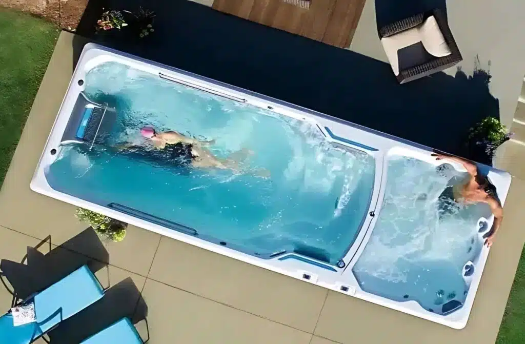 Vue aérienne d'un spa de nage rectangulaire dans un jardin ; une personne nage à gauche, tandis qu'une autre se détend dans le bain bouillonnant à droite. Des chaises de patio et une table se trouvent à proximité.