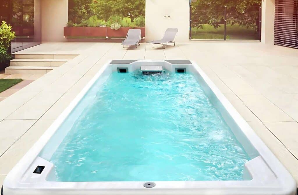 Un jardin moderne comprend une piscine rectangulaire aux eaux bleu clair, entourée de carrelage clair. Deux chaises longues sont installées au fond, près de grandes baies vitrées et d'une végétation luxuriante.