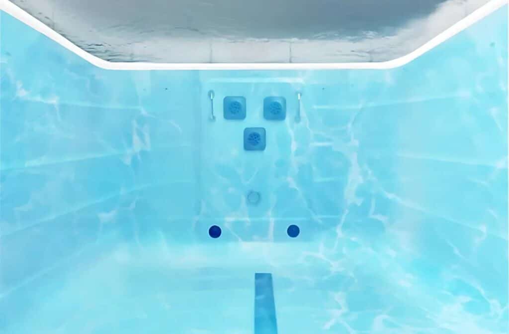 Vue sous-marine d'une piscine aux parois carrelées de bleu, avec deux poignées et des aérations visibles sur le mur du fond. La lumière venant du dessus crée des reflets ondulants sur les surfaces du bassin.
