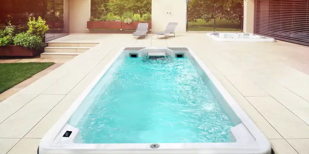 Le Spa de nage D600P, aux eaux cristallines, est installé sur une spacieuse terrasse carrelée. Deux chaises longues et de la verdure se dessinent en arrière-plan, à côté d'un autre jacuzzi près d'une maison moderne.