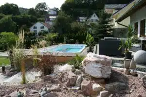 Une cour arrière paysagée comprenant un jacuzzi surélevé entouré d'une terrasse en bois avec mobilier d'extérieur. Le jardin comprend diverses roches décoratives, des plantes et une allée de gravier. En arrière-plan, des maisons et des arbres à flanc de colline sous un ciel dégagé.