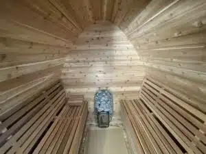 L'image montre l'intérieur d'un sauna en bois avec des plafonds voûtés, des murs et des bancs en bois. Au fond de la pièce se trouve un poêle en pierre utilisé pour produire de la chaleur et de la vapeur. L'atmosphère générale suggère un espace cosy et relaxant.