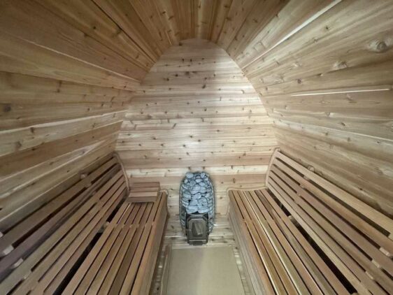 L'image montre l'intérieur d'un sauna en bois avec des plafonds voûtés, des murs et des bancs en bois. Au fond de la pièce se trouve un poêle en pierre utilisé pour produire de la chaleur et de la vapeur. L'atmosphère générale suggère un espace cosy et relaxant.