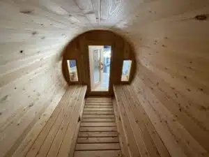 Vue intérieure d'un sauna en tonneau en bois avec deux fenêtres étroites de chaque côté d'une porte en bois à l'extrémité. Les murs, le sol et les bancs sont en bois clair. L'espace est éclairé par la lumière naturelle qui traverse les fenêtres et la porte.