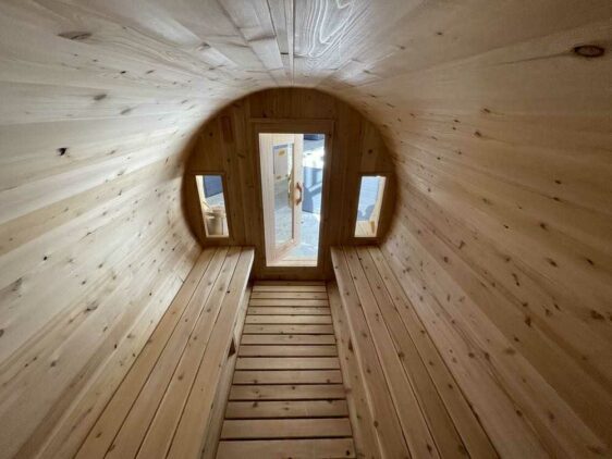 Vue intérieure d'un sauna en tonneau en bois avec deux fenêtres étroites de chaque côté d'une porte en bois à l'extrémité. Les murs, le sol et les bancs sont en bois clair. L'espace est éclairé par la lumière naturelle qui traverse les fenêtres et la porte.