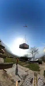 Un hélicoptère hisse un grand objet rectangulaire au-dessus d'une terrasse entourée de zones herbeuses et d'arbres. Une personne portant un gilet de sécurité se tient en dessous et observe l'opération. L’arrière-plan présente une vue panoramique de collines lointaines sous un ciel bleu clair.