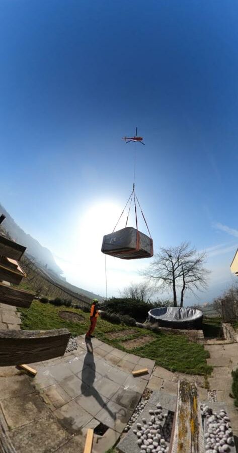 Un hélicoptère hisse un grand objet rectangulaire au-dessus d'une terrasse entourée de zones herbeuses et d'arbres. Une personne portant un gilet de sécurité se tient en dessous et observe l'opération. L’arrière-plan présente une vue panoramique de collines lointaines sous un ciel bleu clair.