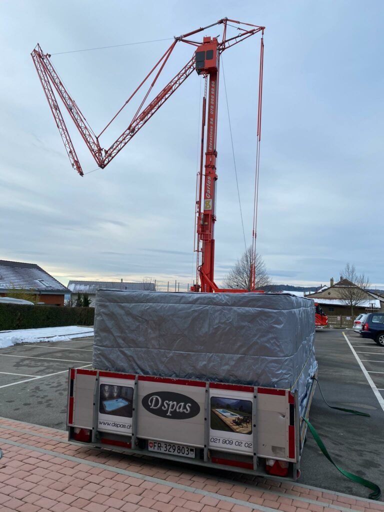 Une grue de chantier rouge est installée sur un parking, à côté d'une remorque recouverte d'une bâche grise. La remorque porte une plaque d'immatriculation indiquant « FR 329803 » et un panneau annonçant « Dspas ». Le ciel est nuageux et la neige est visible au sol et sur les toits.