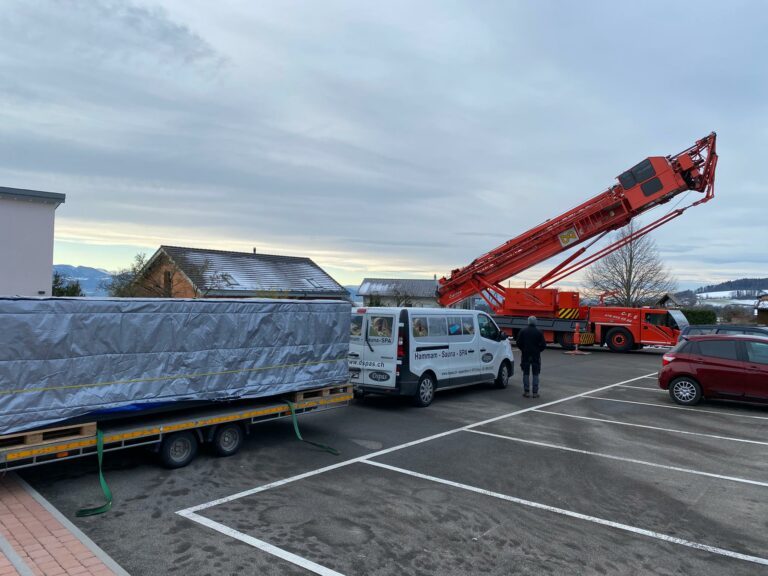 Un gros objet couvert est transporté par une grue et une remorque dans un parking ouvert. On peut voir une camionnette utilitaire blanche avec « Autotrasporti » et une personne à côté. Un ciel couvert et une voiture rouge au premier plan sont visibles. Les environs comprennent des bâtiments et des arbres.