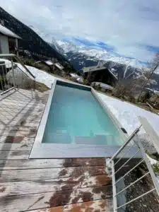 Un bain à remous rectangulaire extérieur étroit avec de l’eau bleu clair est installé contre une terrasse surplombant un flanc de montagne enneigé. Le paysage comprend des maisons dispersées, des arbres et un décor spectaculaire de sommets enneigés sous un ciel partiellement nuageux.