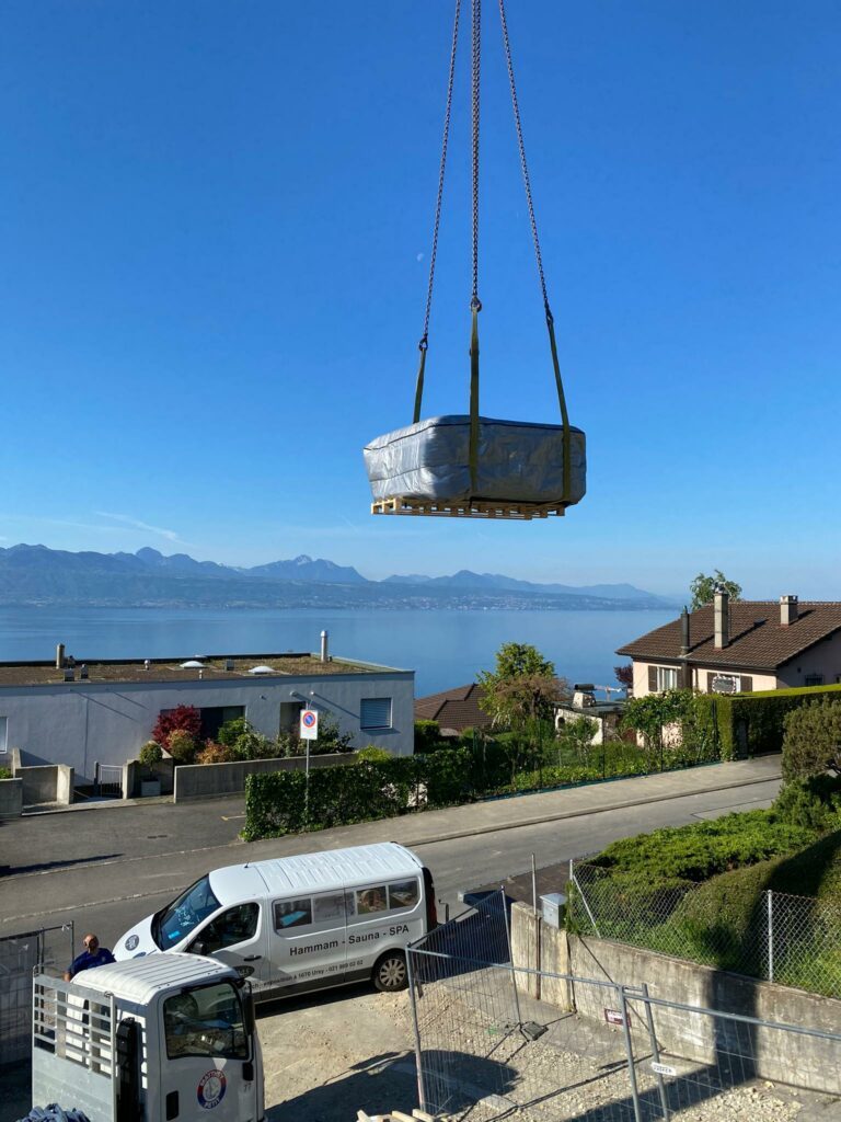 Un grand objet rectangulaire enveloppé dans un matériau de protection est soulevé par une grue au-dessus d'une rue résidentielle. Ci-dessous, une camionnette avec un texte sur le côté est garée dans la rue. En arrière-plan, on a une vue dégagée sur un lac serein et des montagnes sous un ciel bleu.