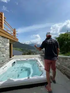 Un homme vêtu d’une chemise noire et d’un short rouge se tient debout sur une terrasse dotée d’un jacuzzi, surplombant une vallée entourée de montagnes. Le ciel est partiellement nuageux et une balustrade en bois est visible sur le côté gauche de la terrasse.
