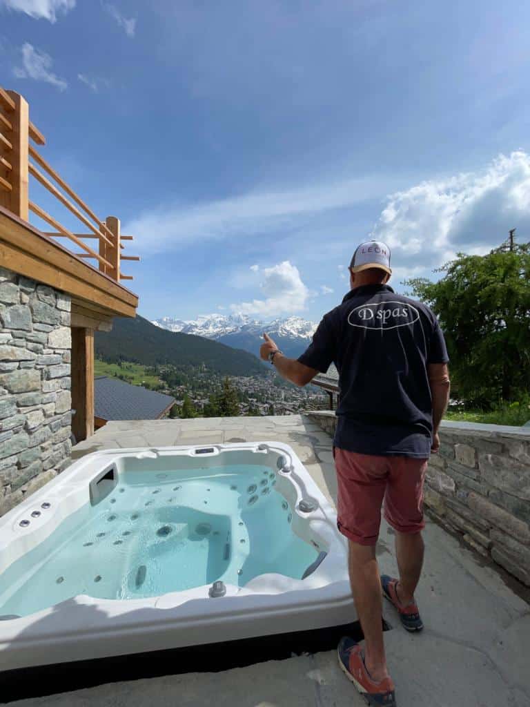 Un homme vêtu d’une chemise noire et d’un short rouge se tient debout sur une terrasse dotée d’un jacuzzi, surplombant une vallée entourée de montagnes. Le ciel est partiellement nuageux et une balustrade en bois est visible sur le côté gauche de la terrasse.