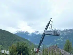Une grue soulève un grand objet couvert au milieu d'un terrain montagneux pittoresque avec des vallées verdoyantes et des sommets partiellement enneigés sous un ciel couvert. Les pins sont au premier plan et les toits en bois sont visibles en contrebas.