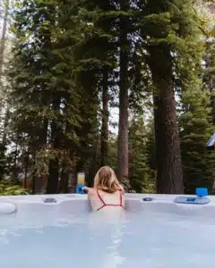 Une personne aux cheveux longs est assise dans un jacuzzi extérieur, vêtue d’un bikini rouge. Ils font face, regardant les grands pins qui entourent la zone, avec une tasse bleue et une boisson dans un verre transparent posés au bord du spa.