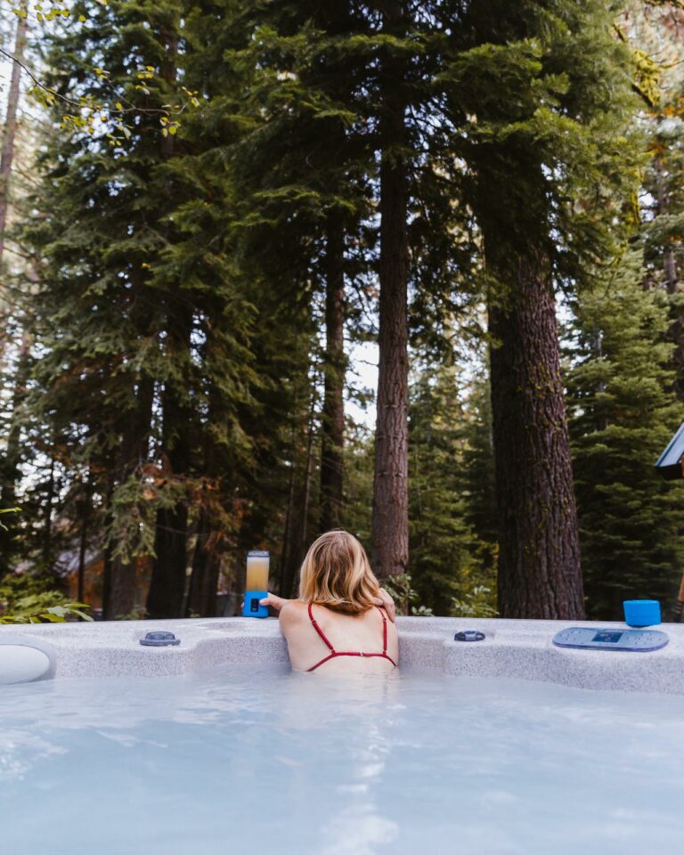 Une personne aux cheveux longs est assise dans un jacuzzi extérieur, vêtue d’un bikini rouge. Ils font face, regardant les grands pins qui entourent la zone, avec une tasse bleue et une boisson dans un verre transparent posés au bord du spa.
