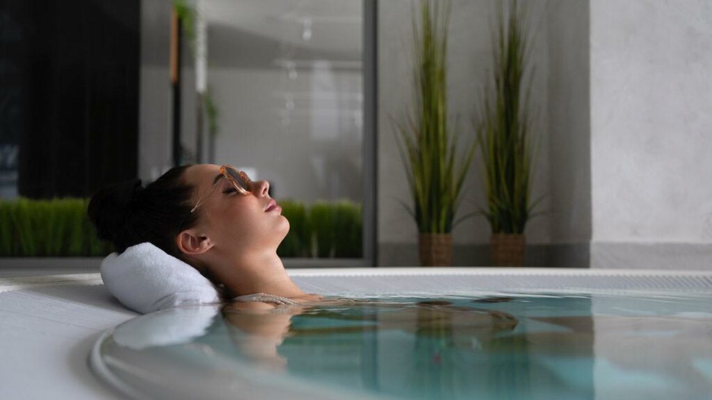 Une femme se détend dans un jacuzzi chromé, la tête posée sur une serviette. Elle est allongée, les yeux fermés et un léger sourire sur le visage. De grandes plantes vertes dans des paniers tressés sont visibles à l'arrière-plan, contribuant au cadre tranquille, semblable à celui d'un spa, qui nécessite un minimum d'entretien.