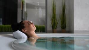 Une femme se détend dans un jacuzzi chromé, la tête posée sur une serviette. Elle est allongée, les yeux fermés et un léger sourire sur le visage. De grandes plantes vertes dans des paniers tressés sont visibles à l'arrière-plan, contribuant au cadre tranquille, semblable à celui d'un spa, qui nécessite un minimum d'entretien.