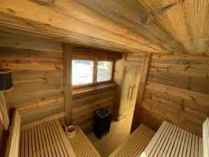 Un sauna confortable en bois avec deux bancs en bois se faisant face, une fenêtre laissant entrer la lumière naturelle et un poêle en pierres. La pièce présente des boiseries rustiques sur les murs et le plafond, lui conférant une atmosphère chaleureuse et accueillante.