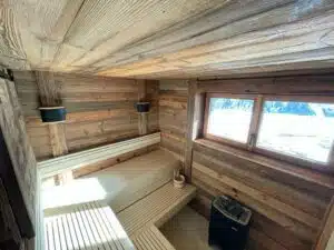 Un sauna en bois confortable avec des murs et des bancs chaleureux en bois. Deux appliques noires se trouvent sur le côté gauche. Une fenêtre à droite laisse entrer la lumière naturelle et offre une vue sur les montagnes enneigées. Le chauffe-sauna se trouve dans le coin inférieur droit.