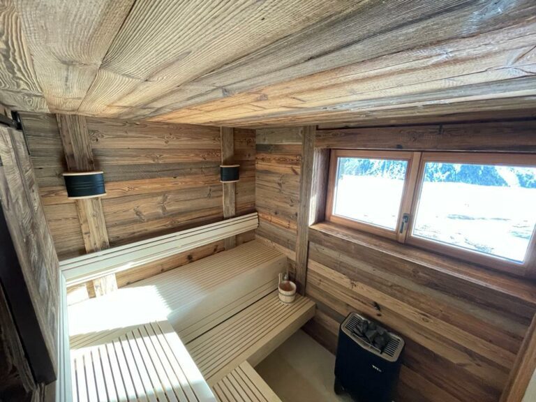 Un sauna en bois confortable avec des murs et des bancs chaleureux en bois. Deux appliques noires se trouvent sur le côté gauche. Une fenêtre à droite laisse entrer la lumière naturelle et offre une vue sur les montagnes enneigées. Le chauffe-sauna se trouve dans le coin inférieur droit.