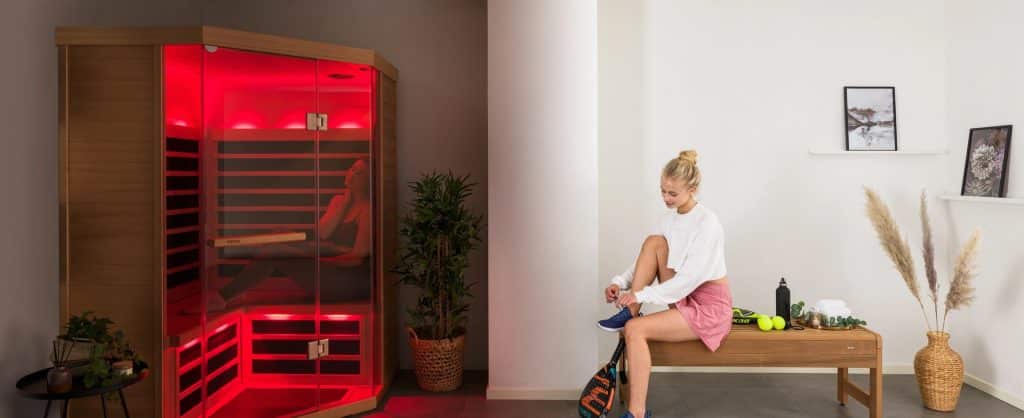 Une femme est assise sur un banc en bois dans un espace bien-être et attache ses chaussures. Derrière elle, une personne se détend dans un sauna infrarouge lambrissé de bois et éclairé en rouge. La pièce est décorée de plantes, de tableaux encadrés et d'une étagère contenant de petits objets.