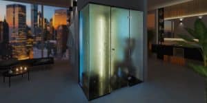 Une salle de bain moderne et faiblement éclairée avec un grand espace douche vitré rappelant un hammam, éclairé de l'intérieur et offrant une vue sur les toits de la ville à travers une large fenêtre. La pièce dispose également d'un lavabo élégant avec un miroir et une décoration de plantes vertes.