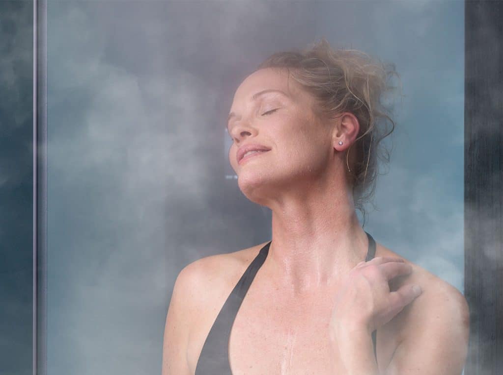 Une personne se tient debout dans le hammam torride, les yeux fermés avec une expression détendue. Ils portent un débardeur noir et leurs cheveux sont tirés en arrière. Ils semblent apprécier la chaleur et la vapeur qui les entourent.