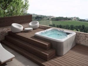 Une terrasse extérieure moderne dotée d'un jacuzzi encastré avec de l'eau claire et bouillonnante. Autour du bain à remous se trouvent d'élégantes chaises longues en osier blanc et une petite table. La terrasse donne sur une vue panoramique sur des vignobles verdoyants et des collines lointaines sous un ciel bleu clair.