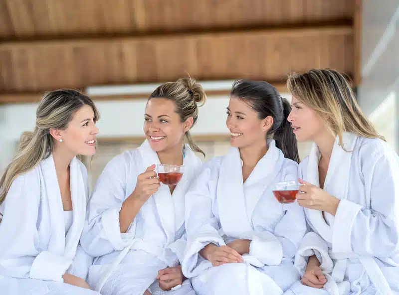 Quatre femmes en robe blanche sont assises ensemble, souriantes et savourant leur tasse de thé. Elles se prélassent dans l'atmosphère apaisante d'un spa, avec des touches de chaleur d'un sauna, des poutres en bois au plafond visibles au-dessus d'elles.