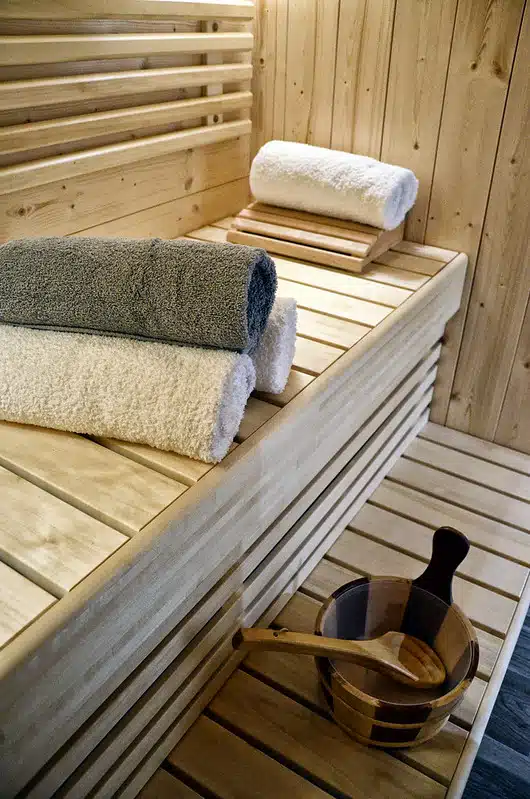 L'intérieur du sauna en bois présente des serviettes soigneusement empilées en gris et blanc sur le banc. Un seau et une louche en bois reposent sur le siège, parfaitement assortis aux panneaux muraux. L'atmosphère du sauna est chaleureuse et accueillante.