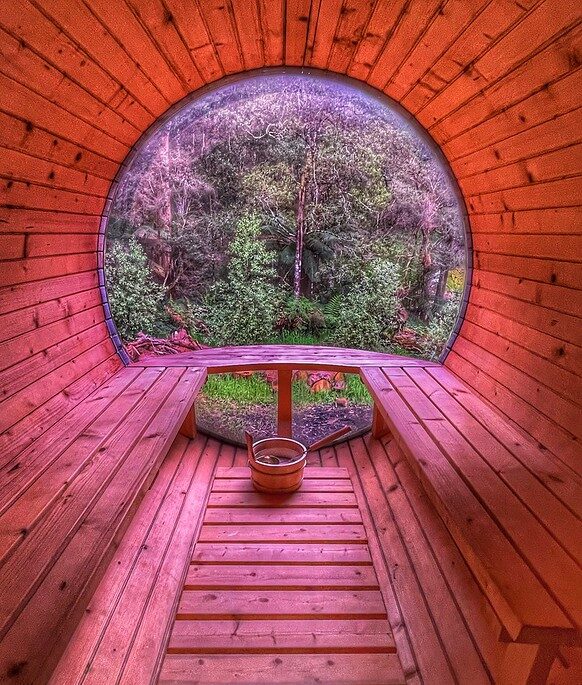 L'intérieur chaleureux du sauna en bois présente une forme cylindrique unique, avec des bancs intégrés sur les côtés. Une grande fenêtre ronde à l'avant encadre une vue sereine sur les arbres et le feuillage verdoyants, renforçant l'atmosphère tranquille de cette retraite de sauna accueillante.