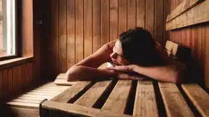 Une personne est allongée sur un banc en bois dans le sauna, la tête appuyée sur ses bras. La pièce est faite de boiseries et la lumière filtre par une fenêtre sur le côté gauche.