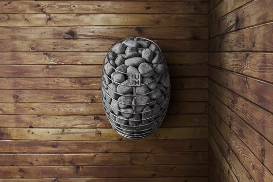 Un chauffe-sauna moderne rempli de pierres grises est monté sur un mur en bois. Le bois est disposé horizontalement, donnant une touche rustique au décor du sauna. Le radiateur a un design cylindrique élégant avec des barres métalliques entourant les pierres. Le nom de la marque « HUUM » est visible.