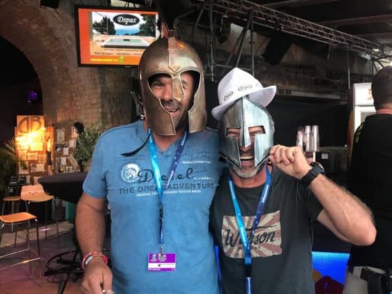 Lors d’un événement animé, deux hommes portent des casques de style médiéval tout en souriant et en posant pour une photo. Tous deux ont des badges d’événement et des cordons autour du cou. L’un porte un t-shirt avec du texte et l’autre ajoute un chapeau de cowboy au sommet de son casque. L’arrière-plan montre le décor et les écrans de l’événement. membres de Dspas
