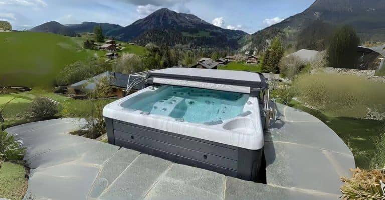 Un bain à remous avec de l'eau bouillonnante est installé sur une terrasse en pierre, surplombant une vallée pittoresque avec des collines verdoyantes, des maisons dispersées et des montagnes imposantes sous un ciel partiellement nuageux.