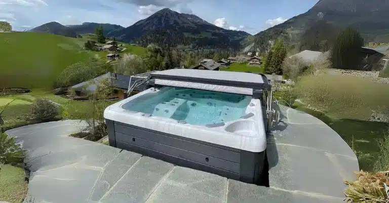 Un bain à remous avec de l'eau bouillonnante est installé sur une terrasse en pierre, surplombant une vallée pittoresque avec des collines verdoyantes, des maisons dispersées et des montagnes imposantes sous un ciel partiellement nuageux.