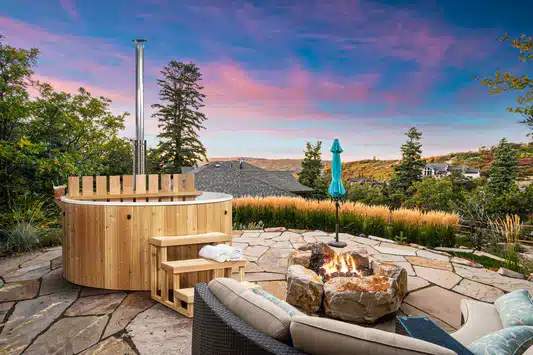 Un patio extérieur confortable comprend le jacuzzi Bain nordique en bois, un foyer en pierre, des sièges rembourrés et un parasol bleu fermé, le tout niché au milieu des arbres et des maisons sous un ciel de couchers de soleil éclatants.