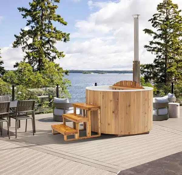 Un Bain nordique en bois avec marches se trouve sur une terrasse au bord d'un lac, entouré de conifères. Des chaises d'extérieur et une table à manger à proximité en font l'endroit idéal pour se détendre et se relaxer sous un ciel partiellement nuageux.