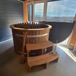 Le Bain nordique en bois avec bandes métalliques est installé à l'intérieur, près de grandes fenêtres, avec des marches en bois qui y mènent, une pagaie à proximité, et son couvercle isolant noir replié derrière la baignoire.