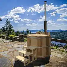 Le Bain nordique en bois avec une cheminée en métal se trouve sur un patio en pierre, offrant une vue sur les arbres, les collines et un lac sous un ciel bleu vif avec des nuages épars.