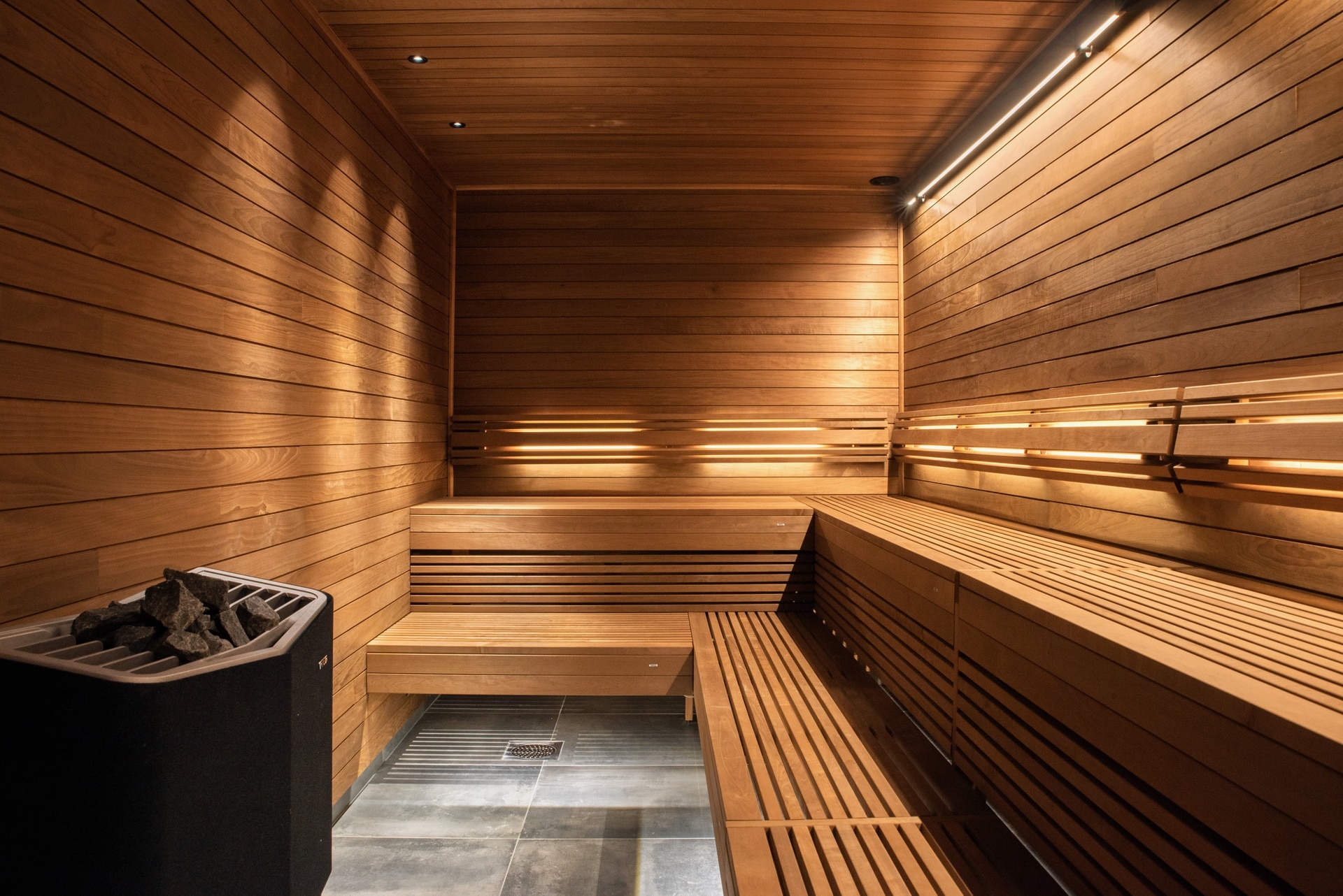 Un sauna chaleureux et minimaliste avec des bancs et des murs en bois Tylo. Un éclairage tamisé crée une ambiance relaxante, tandis qu'un poêle à pierres est visible dans le coin. Le sol carrelé complète le design élégant en bois.