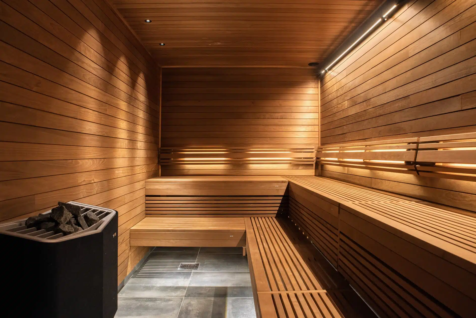 Un sauna chaleureux et minimaliste avec des bancs et des murs en bois Tylo. Un éclairage tamisé crée une ambiance relaxante, tandis qu'un poêle à pierres est visible dans le coin. Le sol carrelé complète le design élégant en bois.