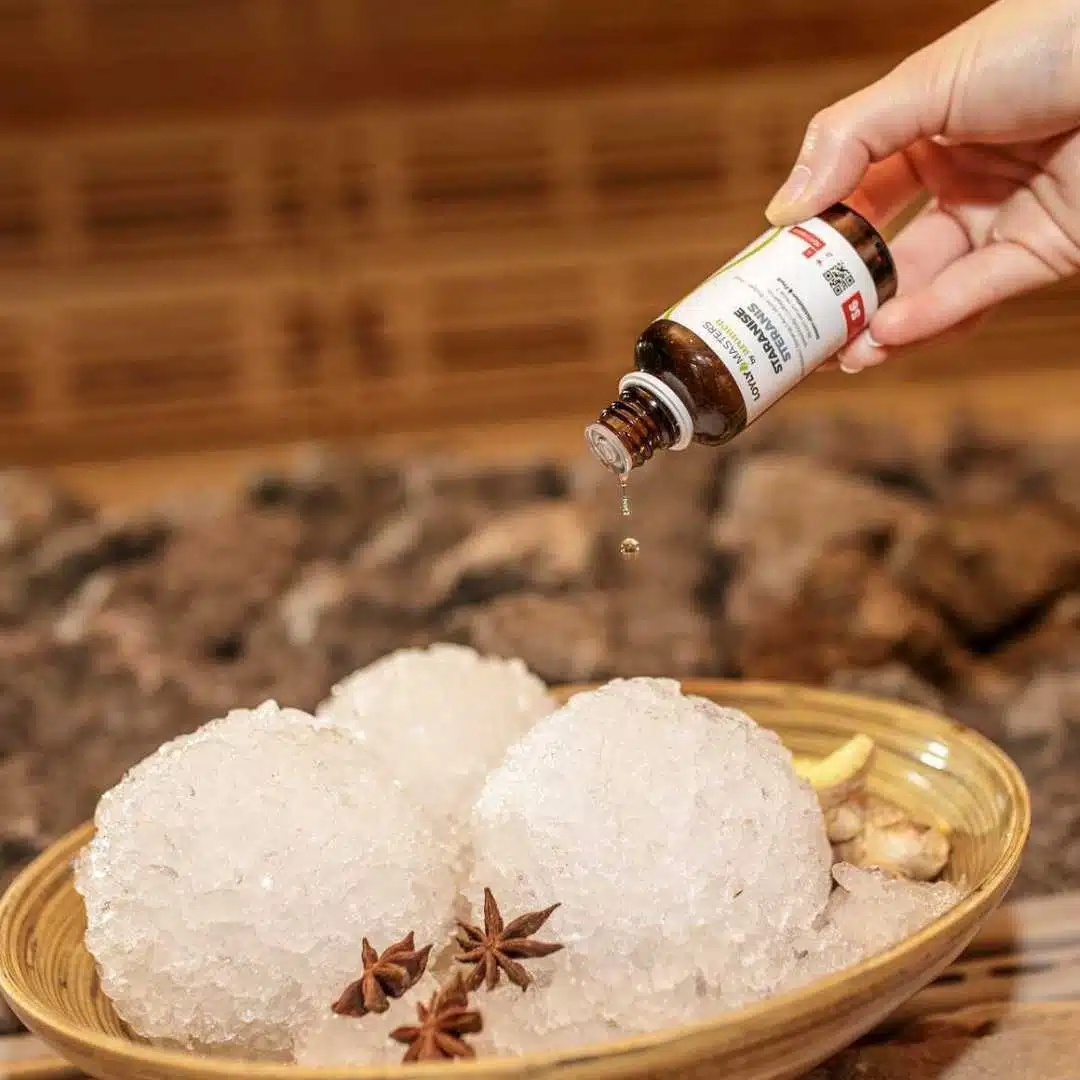 Une main dépose des gouttes d'une petite bouteille brune sur trois gros amas de glace sphériques dans un bol, rappelant l'ambiance sereine d'un bain nordique en Suisse. La glace est agrémentée de tranches d'anis étoilé et de gingembre, le tout sur un fond texturé rustique.
