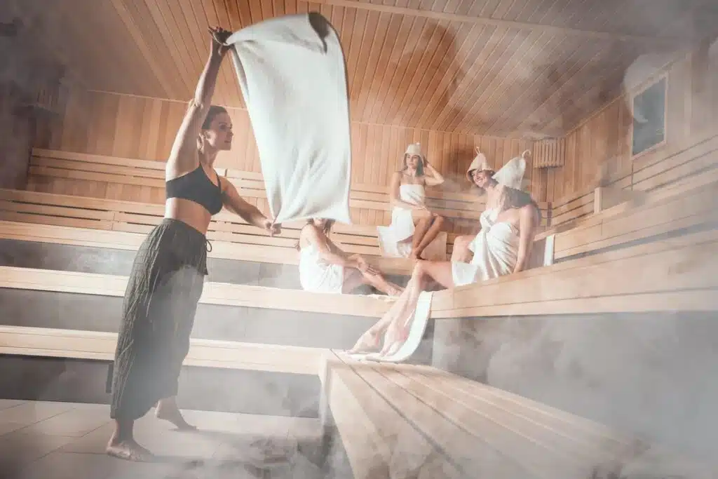 Une femme vêtue d'un haut de sport noir et d'un pantalon sombre agite une serviette dans un sauna fumant, rappelant un bain nordique en Suisse. Quatre personnes enveloppées dans des serviettes et portant des couvre-chefs sont assises sur des bancs en bois, se relaxant dans l'atmosphère brumeuse.
