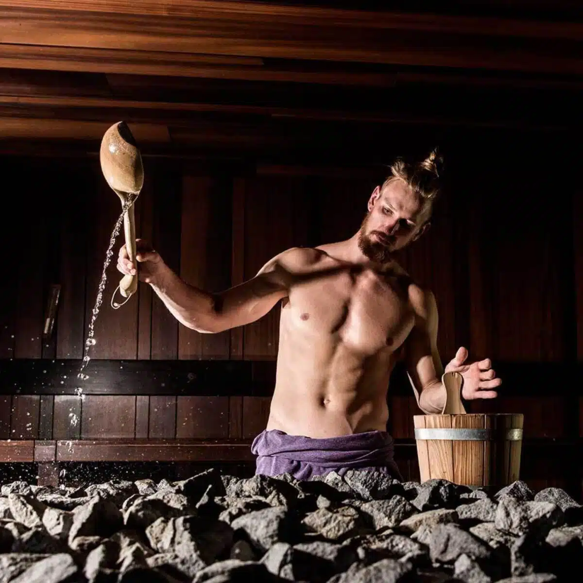 Un homme torse nu, barbu et coiffé d'un chignon, verse de l'eau à l'aide d'une louche en bois sur des pierres chauffées dans un sauna faiblement éclairé aux murs et au plafond en bois. Debout à côté d'un seau en bois, vêtu d'une serviette violette, il évoque l'essence tranquille du bain nordique en Suisse.