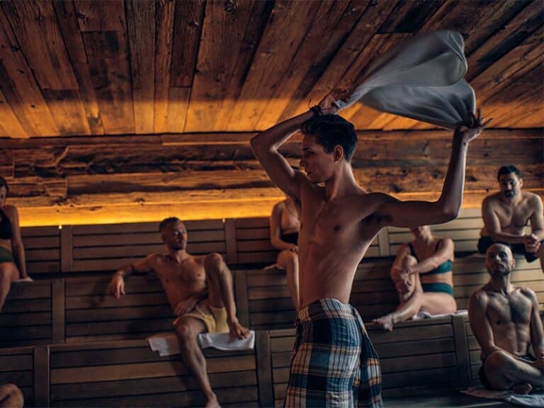 Une personne vêtue d'une serviette à carreaux agite un torchon dans un sauna en bois confortable, rappelant un bain nordique en Suisse. Plusieurs personnes sont assises sur des bancs en bois, certaines avec des serviettes. L'espace est doté d'un éclairage chaleureux et d'une atmosphère rustique.
