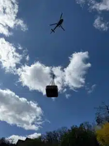 Un hélicoptère survole le paysage, se découpant sur un ciel bleu partiellement nuageux, transportant un grand conteneur suspendu à un câble. Des arbres et des arbustes sont visibles au bas de l'image.