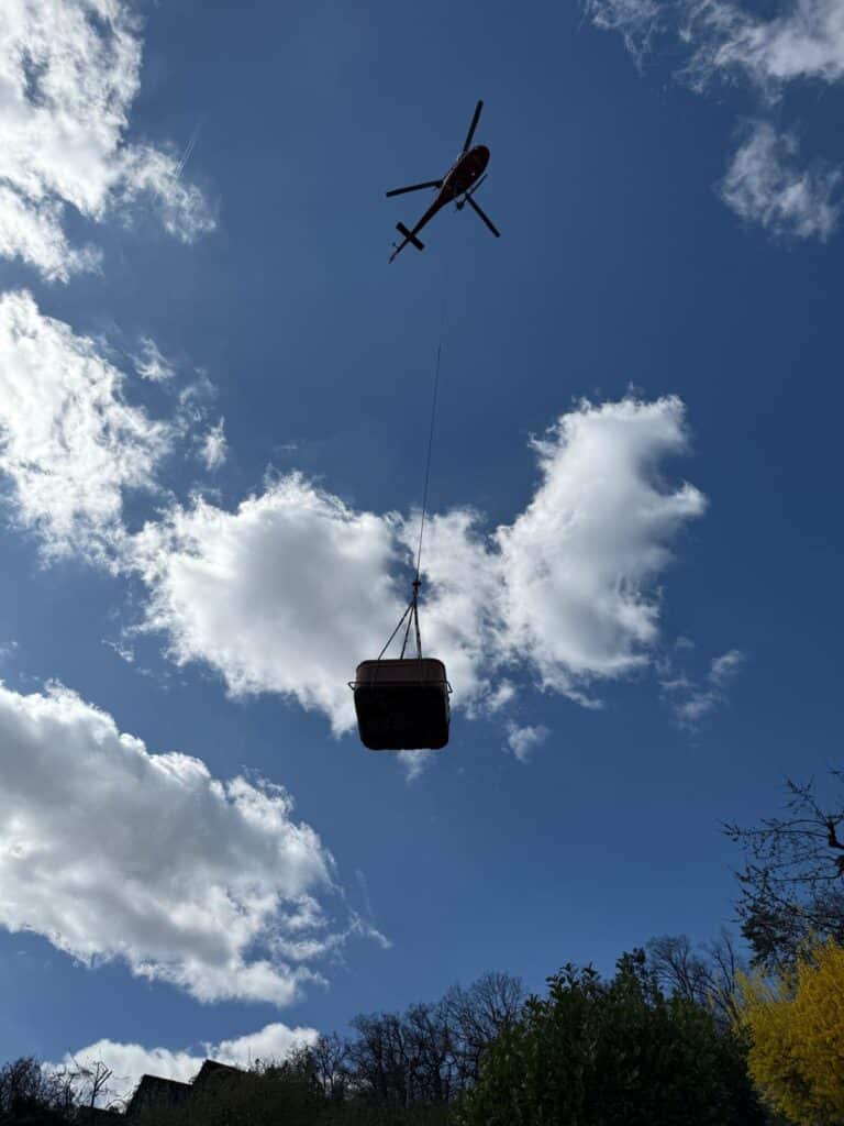 Un hélicoptère survole le paysage, se découpant sur un ciel bleu partiellement nuageux, transportant un grand conteneur suspendu à un câble. Des arbres et des arbustes sont visibles au bas de l'image.